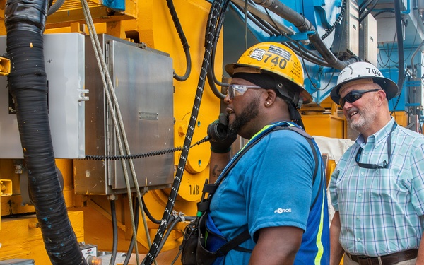 Two workers are standing beside large yellow industrial machinery at what appears to be a port or construction site. One worker wearing a yellow hard hat and blue safety vest is speaking on a corded phone attached to the equipment, while a second worker in a white hard hat and plaid shirt looks on smiling. A crane and port infrastructure are visible in the background.