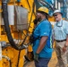 Two workers are standing beside large yellow industrial machinery at what appears to be a port or construction site. One worker wearing a yellow hard hat and blue safety vest is speaking on a corded phone attached to the equipment, while a second worker in a white hard hat and plaid shirt looks on smiling. A crane and port infrastructure are visible in the background.