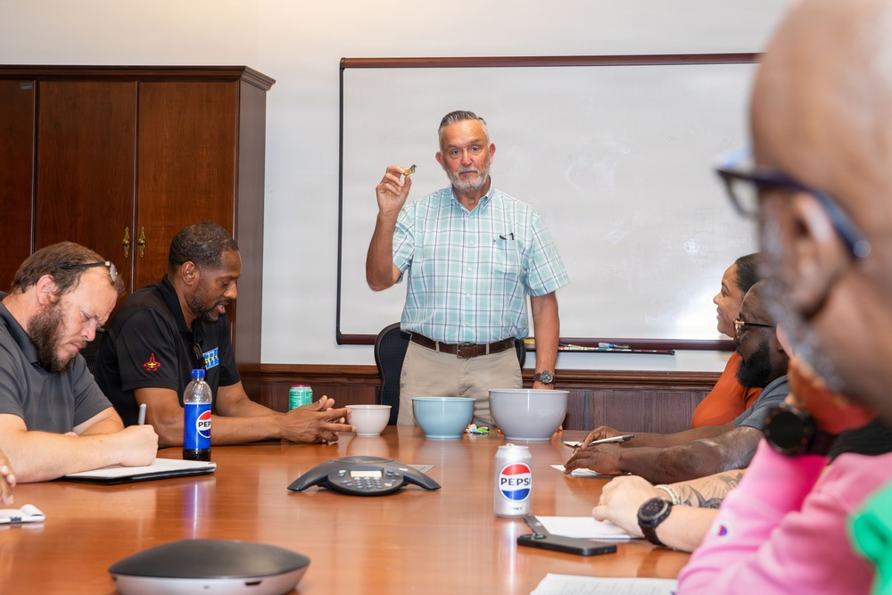 A man in a light plaid shirt stands at the head of a conference table, holding up a small object while presenting to a group of seated attendees. The meeting takes place in a formal boardroom setting with a whiteboard behind the presenter. Several participants have notepads, beverages, and bowls on the table in front of them.
