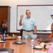 A man in a light plaid shirt stands at the head of a conference table, holding up a small object while presenting to a group of seated attendees. The meeting takes place in a formal boardroom setting with a whiteboard behind the presenter. Several participants have notepads, beverages, and bowls on the table in front of them.