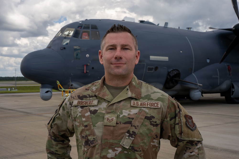A U.S. Air Force Staff Sergeant named Boisvert poses in front of a large military transport aircraft on a flight line. He is wearing OCP (Operational Camouflage Pattern) uniform with visible name tape, branch tape, and rank insignia. The aircraft behind him appears to be a dark gray C-130 variant.