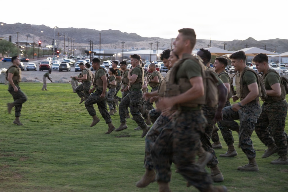 A group of U.S. Marines in camouflage uniforms and tactical vests are running across a grassy field during what appears to be a physical training exercise at dusk or dawn. The setting appears to be a military installation in a desert environment, identifiable by the arid mountains in the background and the base infrastructure visible behind them. Several personnel appear to be smiling and in good spirits during the activity.