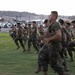 A group of U.S. Marines in camouflage uniforms and tactical vests are running across a grassy field during what appears to be a physical training exercise at dusk or dawn. The setting appears to be a military installation in a desert environment, identifiable by the arid mountains in the background and the base infrastructure visible behind them. Several personnel appear to be smiling and in good spirits during the activity.