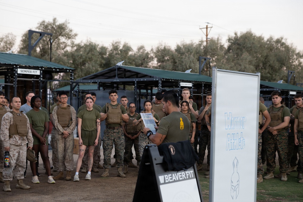 A group of military personnel, likely U.S. Marines based on their uniforms and the 'Semper Fit' signage, are gathered outdoors at a fitness facility for a briefing. An instructor or leader is addressing the group while holding documents, standing next to a sign board reading 'Warrior Athlete' and a 'BeaverFit' branded A-frame sign. The setting appears to be an outdoor functional fitness area with metal rack structures.