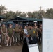 A group of military personnel, likely U.S. Marines based on their uniforms and the 'Semper Fit' signage, are gathered outdoors at a fitness facility for a briefing. An instructor or leader is addressing the group while holding documents, standing next to a sign board reading 'Warrior Athlete' and a 'BeaverFit' branded A-frame sign. The setting appears to be an outdoor functional fitness area with metal rack structures.