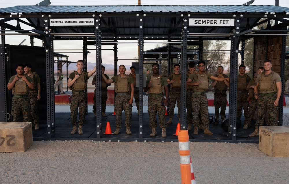 A group of approximately 14 U.S. Marines in combat uniforms and tactical vests are posed together under an outdoor fitness structure. The structure is branded 'American Standard Equipment' and 'Semper Fit,' indicating a military physical training facility. The setting appears to be a desert environment, consistent with a U.S. Marine Corps base.