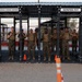 A group of approximately 14 U.S. Marines in combat uniforms and tactical vests are posed together under an outdoor fitness structure. The structure is branded 'American Standard Equipment' and 'Semper Fit,' indicating a military physical training facility. The setting appears to be a desert environment, consistent with a U.S. Marine Corps base.