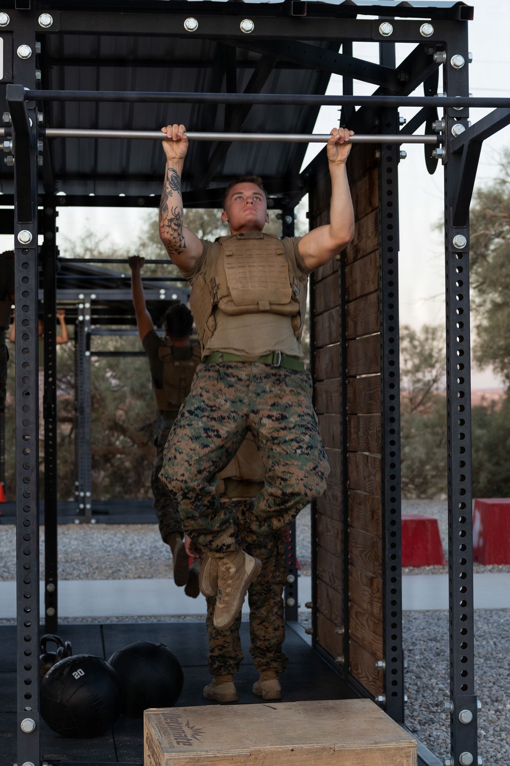 A military service member in Marine Corps MARPAT camouflage uniform and tactical plate carrier vest performs pull-ups on an outdoor fitness rig. At least one additional service member is visible in the background performing the same exercise. The setting appears to be an outdoor military physical training area with kettlebells and a plyo box visible on the ground.