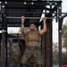 A military service member in Marine Corps MARPAT camouflage uniform and tactical plate carrier vest performs pull-ups on an outdoor fitness rig. At least one additional service member is visible in the background performing the same exercise. The setting appears to be an outdoor military physical training area with kettlebells and a plyo box visible on the ground.
