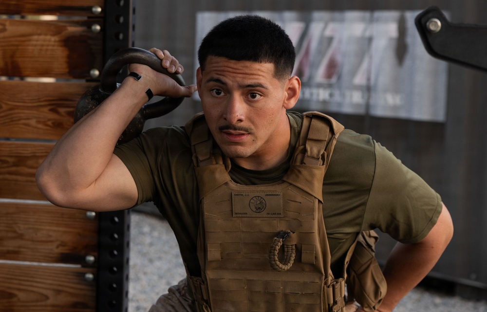 A young male in military or tactical fitness attire is resting against a wooden rack structure in what appears to be a gym or training facility. He is wearing a tan plate carrier vest over an olive green t-shirt and holding a kettlebell handle near his head. His expression appears focused or fatigued, suggesting he is mid-workout.