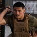 A young male in military or tactical fitness attire is resting against a wooden rack structure in what appears to be a gym or training facility. He is wearing a tan plate carrier vest over an olive green t-shirt and holding a kettlebell handle near his head. His expression appears focused or fatigued, suggesting he is mid-workout.