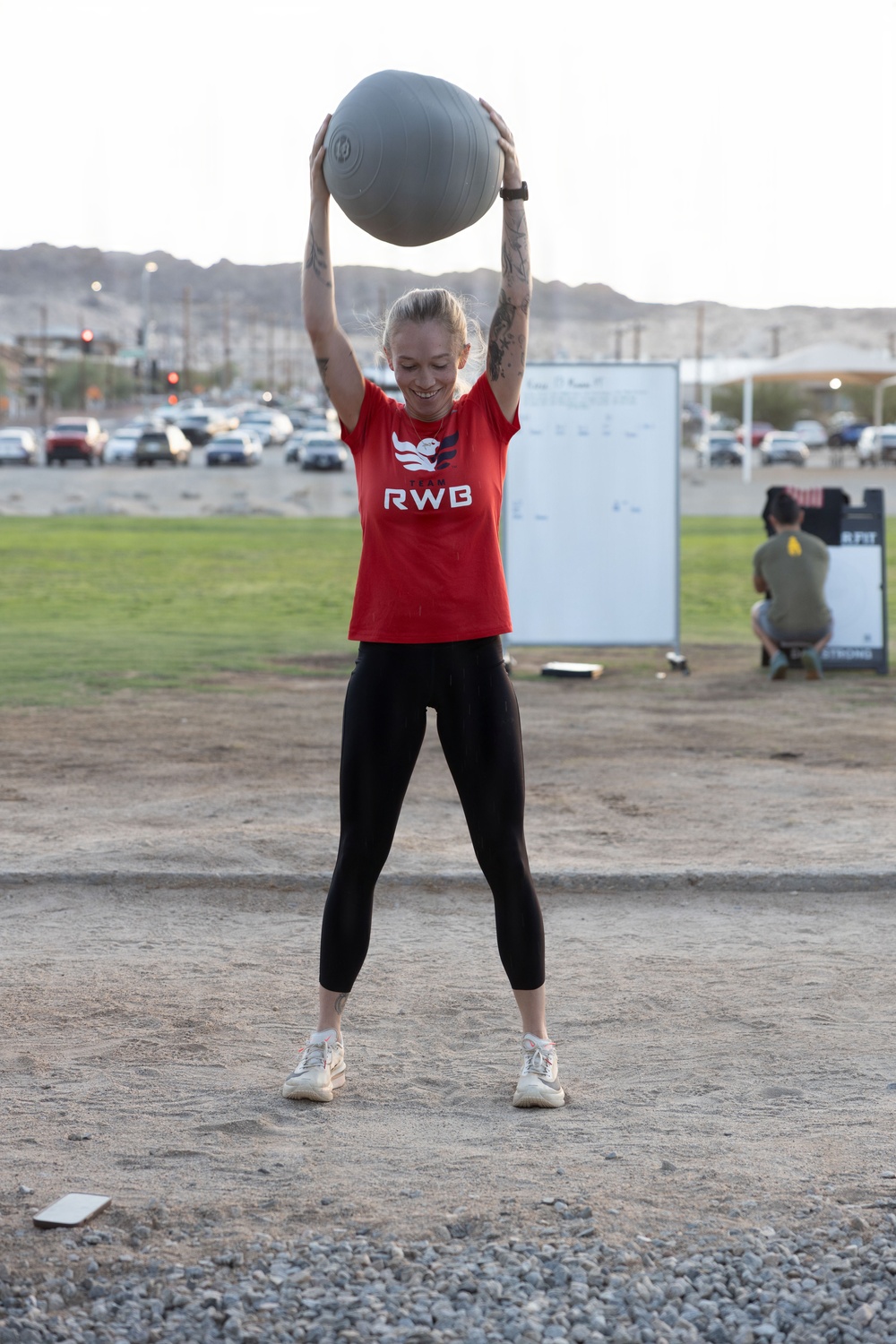 A woman wearing a red 'Team RWB' shirt is performing an overhead medicine ball exercise outdoors on a sandy/gravel surface. She is smiling and has both arms fully extended above her head holding a gray medicine ball. The setting appears to be an outdoor fitness area near a parking lot with desert mountains visible in the background.