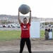 A woman wearing a red 'Team RWB' shirt is performing an overhead medicine ball exercise outdoors on a sandy/gravel surface. She is smiling and has both arms fully extended above her head holding a gray medicine ball. The setting appears to be an outdoor fitness area near a parking lot with desert mountains visible in the background.