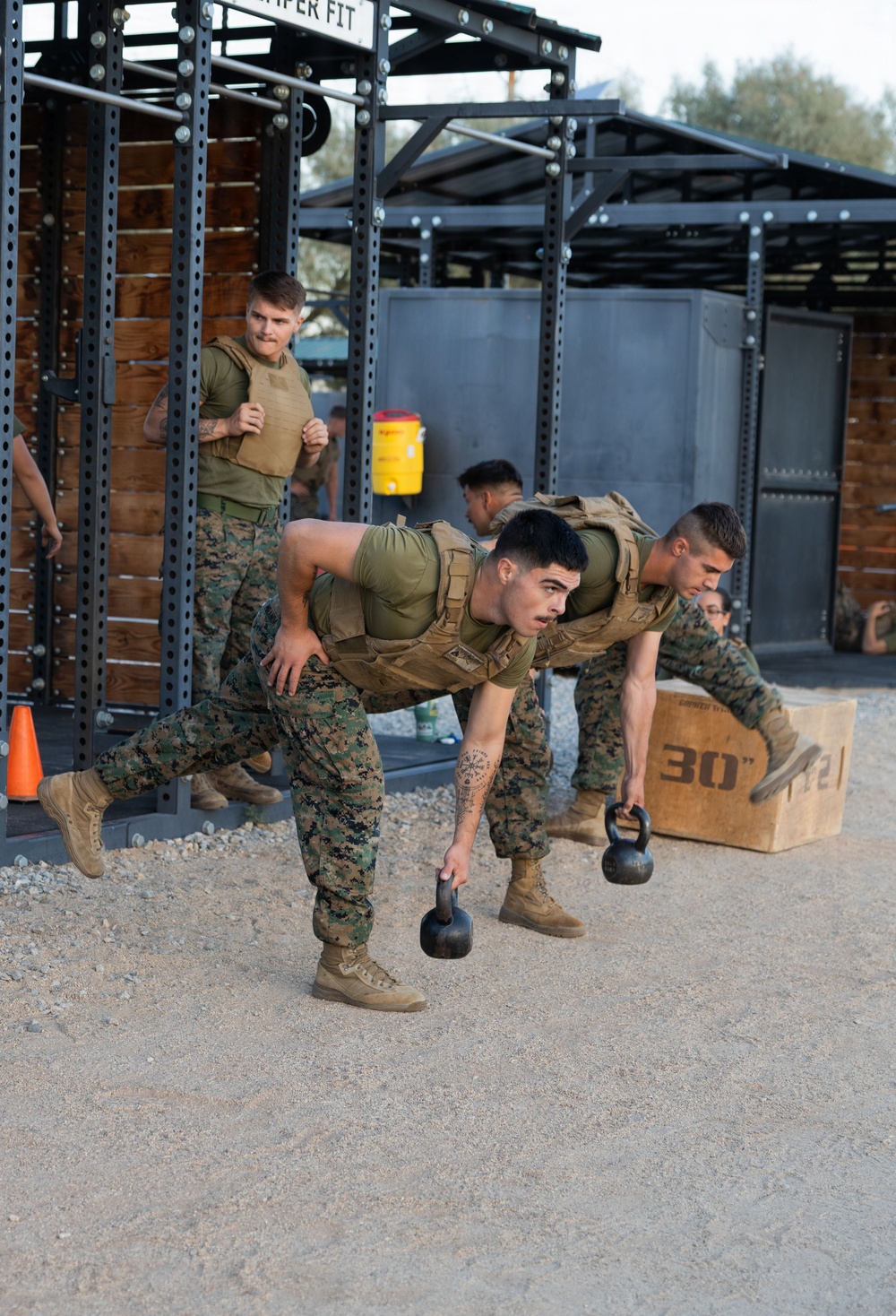 Military personnel in MARPAT camouflage uniforms and tactical vests are conducting a physical training session at an outdoor fitness facility. The foreground shows two service members performing kettlebell exercises in a bent-over position, while a third runs in the background. The setting appears to be a dedicated military fitness area with metal rig structures and gravel ground.