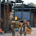 Military personnel in MARPAT camouflage uniforms and tactical vests are conducting a physical training session at an outdoor fitness facility. The foreground shows two service members performing kettlebell exercises in a bent-over position, while a third runs in the background. The setting appears to be a dedicated military fitness area with metal rig structures and gravel ground.
