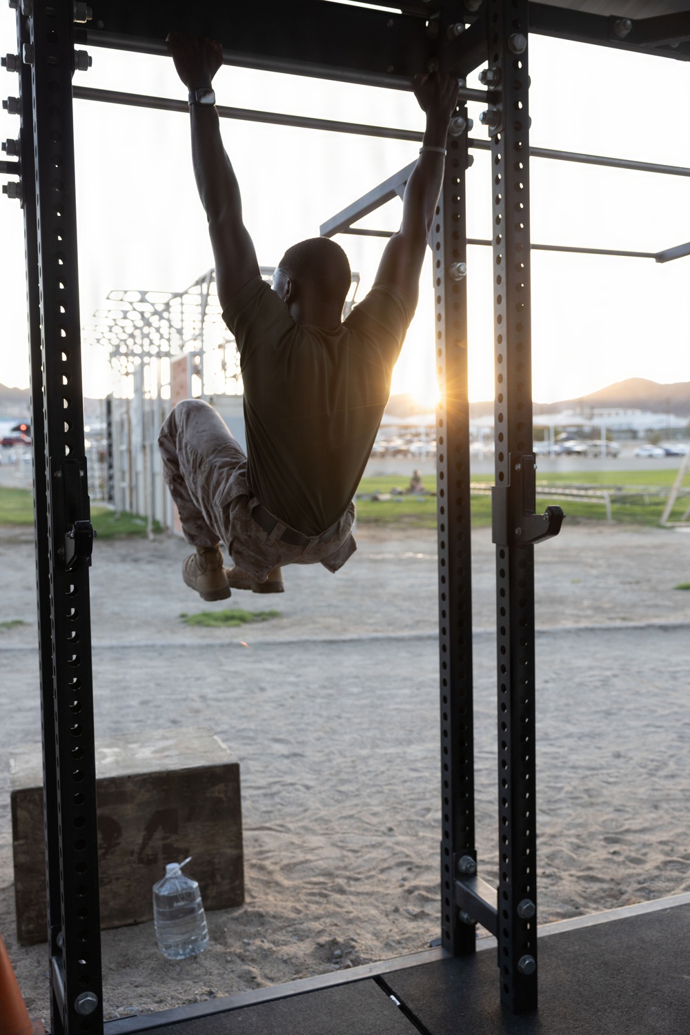 A person in military-style camouflage pants and a dark t-shirt performs a pull-up or muscle-up on an outdoor power rack/pull-up station. The image is backlit by a setting or rising sun, creating a dramatic silhouette effect. The setting appears to be a military or outdoor fitness training area with sandy ground.