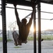 A person in military-style camouflage pants and a dark t-shirt performs a pull-up or muscle-up on an outdoor power rack/pull-up station. The image is backlit by a setting or rising sun, creating a dramatic silhouette effect. The setting appears to be a military or outdoor fitness training area with sandy ground.