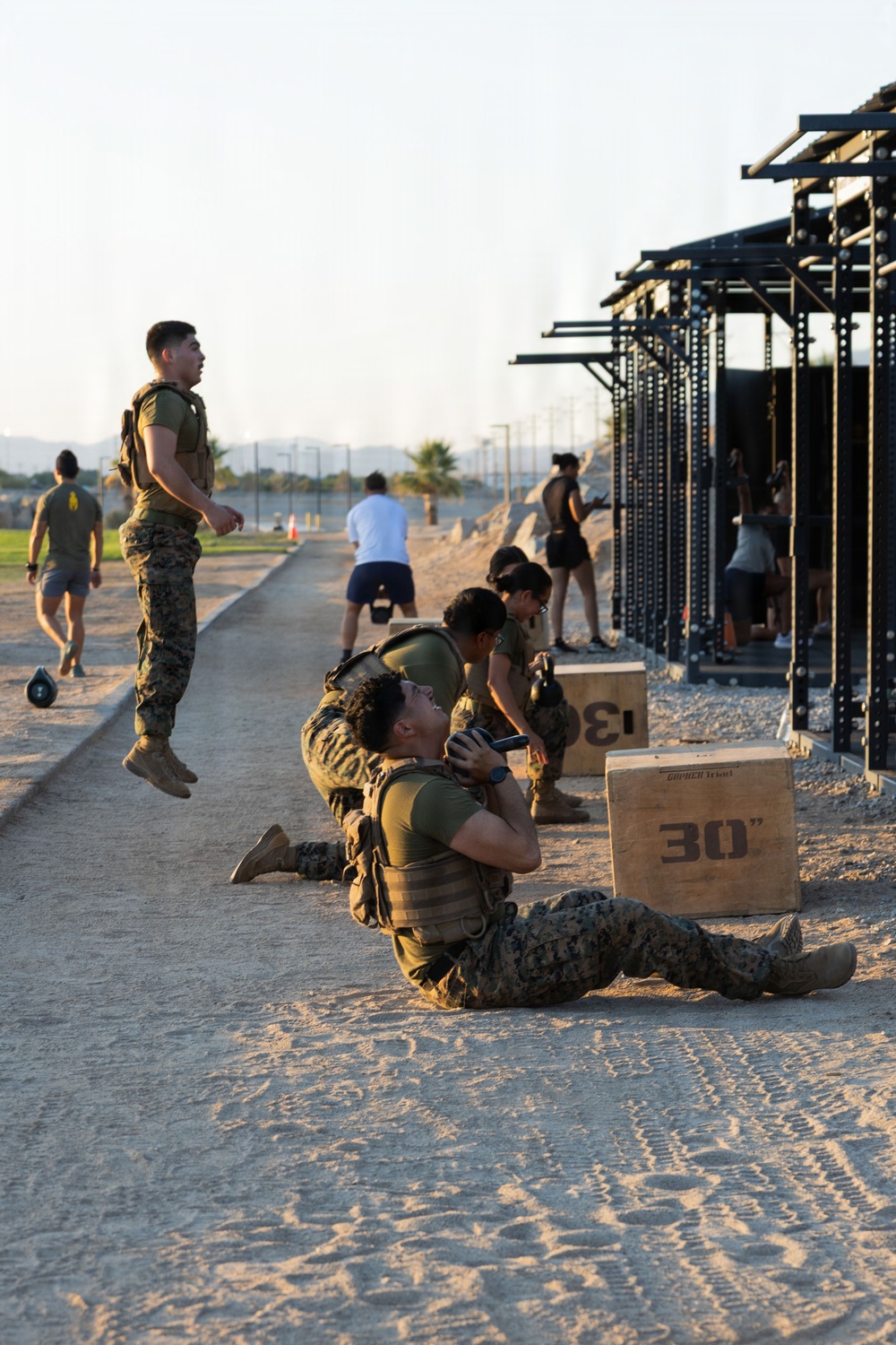 Military personnel in USMC digital camouflage uniforms and tactical vests are conducting a physical training session at an outdoor fitness facility. Multiple exercises are visible simultaneously, including sit-ups, kettlebell work, and jumping. The setting appears to be a military base fitness area with plyometric boxes and a pull-up/rig structure.