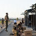 Military personnel in USMC digital camouflage uniforms and tactical vests are conducting a physical training session at an outdoor fitness facility. Multiple exercises are visible simultaneously, including sit-ups, kettlebell work, and jumping. The setting appears to be a military base fitness area with plyometric boxes and a pull-up/rig structure.