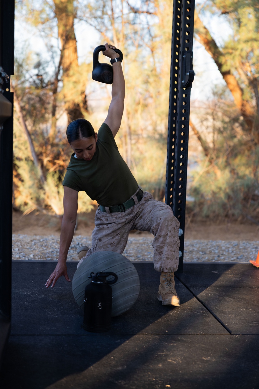 A military service member in USMC desert camouflage utilities and olive drab t-shirt performs a kettlebell Turkish get-up exercise on an outdoor rubber mat platform. One arm is extended overhead holding a black kettlebell while the opposite hand touches the ground for support, with a stability ball positioned beneath the torso. The setting appears to be an outdoor fitness area with a power rack visible and trees in the background.