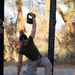 A military service member in USMC desert camouflage utilities and olive drab t-shirt performs a kettlebell Turkish get-up exercise on an outdoor rubber mat platform. One arm is extended overhead holding a black kettlebell while the opposite hand touches the ground for support, with a stability ball positioned beneath the torso. The setting appears to be an outdoor fitness area with a power rack visible and trees in the background.