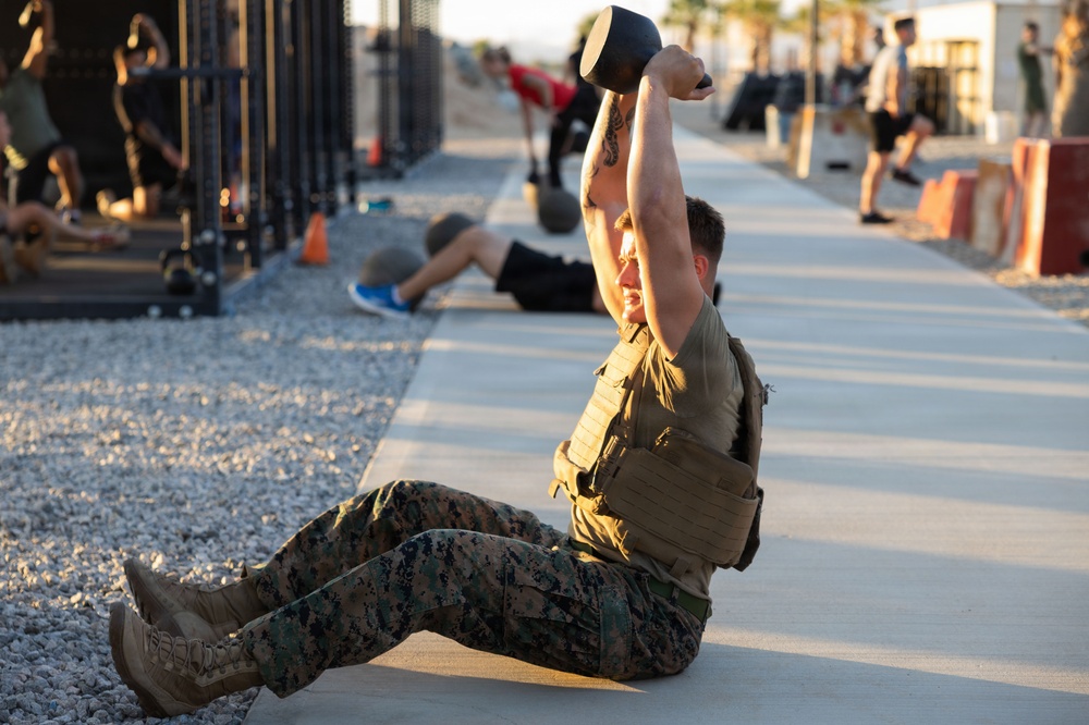 A military service member in USMC digital camouflage (MARPAT) uniform and tactical vest performs a kettlebell sit-up exercise on a concrete surface at an outdoor fitness area. The foreground subject has a visible arm tattoo and is mid-movement, pressing a kettlebell overhead while completing a sit-up. Multiple other personnel are visible in the background engaged in various fitness activities at what appears to be a military base gym or training area.