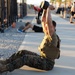 A military service member in USMC digital camouflage (MARPAT) uniform and tactical vest performs a kettlebell sit-up exercise on a concrete surface at an outdoor fitness area. The foreground subject has a visible arm tattoo and is mid-movement, pressing a kettlebell overhead while completing a sit-up. Multiple other personnel are visible in the background engaged in various fitness activities at what appears to be a military base gym or training area.
