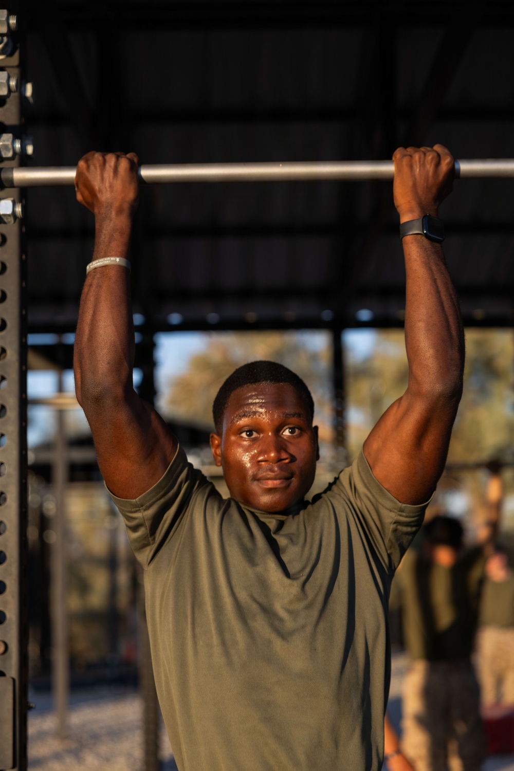A young man is performing pull-ups or hanging from a metal pull-up bar at an outdoor fitness station. He is wearing an olive/army green t-shirt and appears to be sweating, suggesting active physical exertion. The background shows a blurred outdoor environment with other individuals visible in the distance.