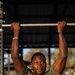A young man is performing pull-ups or hanging from a metal pull-up bar at an outdoor fitness station. He is wearing an olive/army green t-shirt and appears to be sweating, suggesting active physical exertion. The background shows a blurred outdoor environment with other individuals visible in the distance.