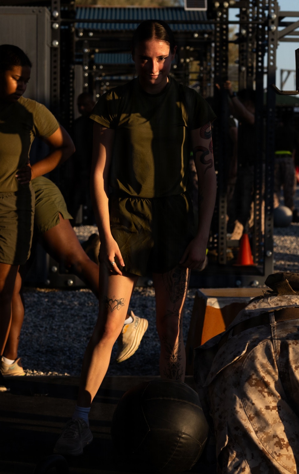 The image shows individuals in olive drab military physical training (PT) uniforms at an outdoor fitness area with power rack equipment visible in the background. The central figure is a young woman standing near what appears to be a medicine ball and military camouflage gear on the ground. The setting appears to be a military base fitness course photographed in warm, low-angle sunlight.