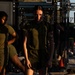 The image shows individuals in olive drab military physical training (PT) uniforms at an outdoor fitness area with power rack equipment visible in the background. The central figure is a young woman standing near what appears to be a medicine ball and military camouflage gear on the ground. The setting appears to be a military base fitness course photographed in warm, low-angle sunlight.