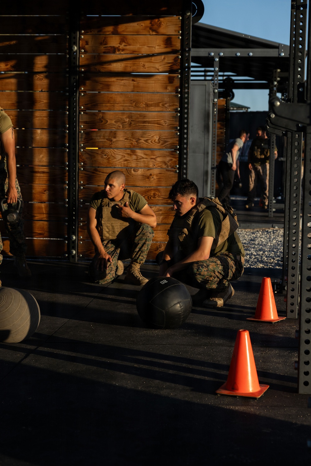 Two military personnel in camouflage uniforms and tactical vests are resting or taking a break at an outdoor fitness facility. They are seated near a wooden wall structure with metal rack frames, surrounded by exercise equipment. Additional personnel are visible in the background of what appears to be a military physical training area.
