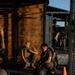 Two military personnel in camouflage uniforms and tactical vests are resting or taking a break at an outdoor fitness facility. They are seated near a wooden wall structure with metal rack frames, surrounded by exercise equipment. Additional personnel are visible in the background of what appears to be a military physical training area.