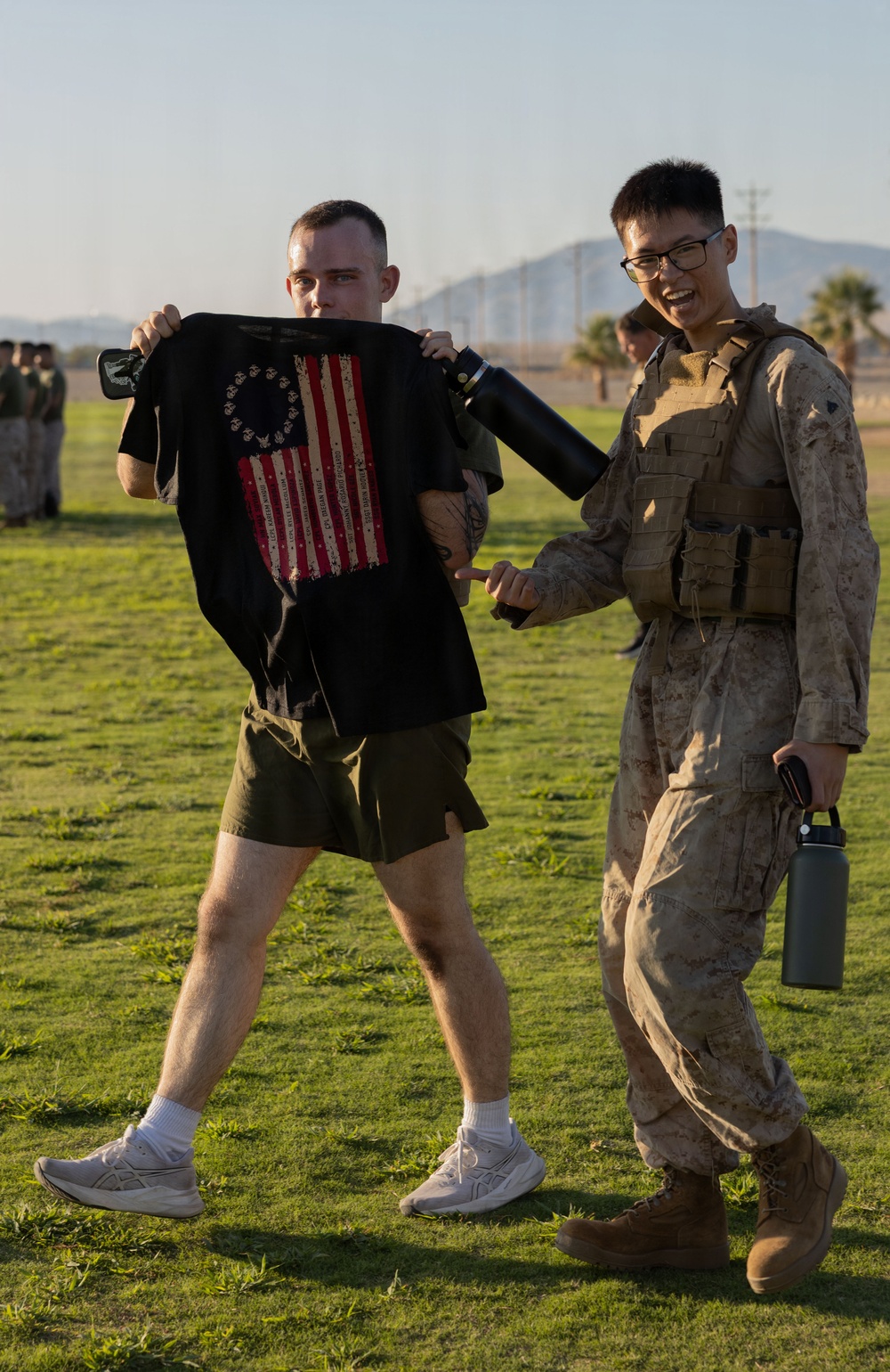 Two individuals stand on a grass field, with one holding up a black t-shirt featuring an American Betsy Ross flag design. The person on the right is wearing Marine Corps MARPAT camouflage utilities with a plate carrier vest and carrying water bottles. A formation of personnel in military attire is visible in the background.