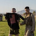 Two individuals stand on a grass field, with one holding up a black t-shirt featuring an American Betsy Ross flag design. The person on the right is wearing Marine Corps MARPAT camouflage utilities with a plate carrier vest and carrying water bottles. A formation of personnel in military attire is visible in the background.