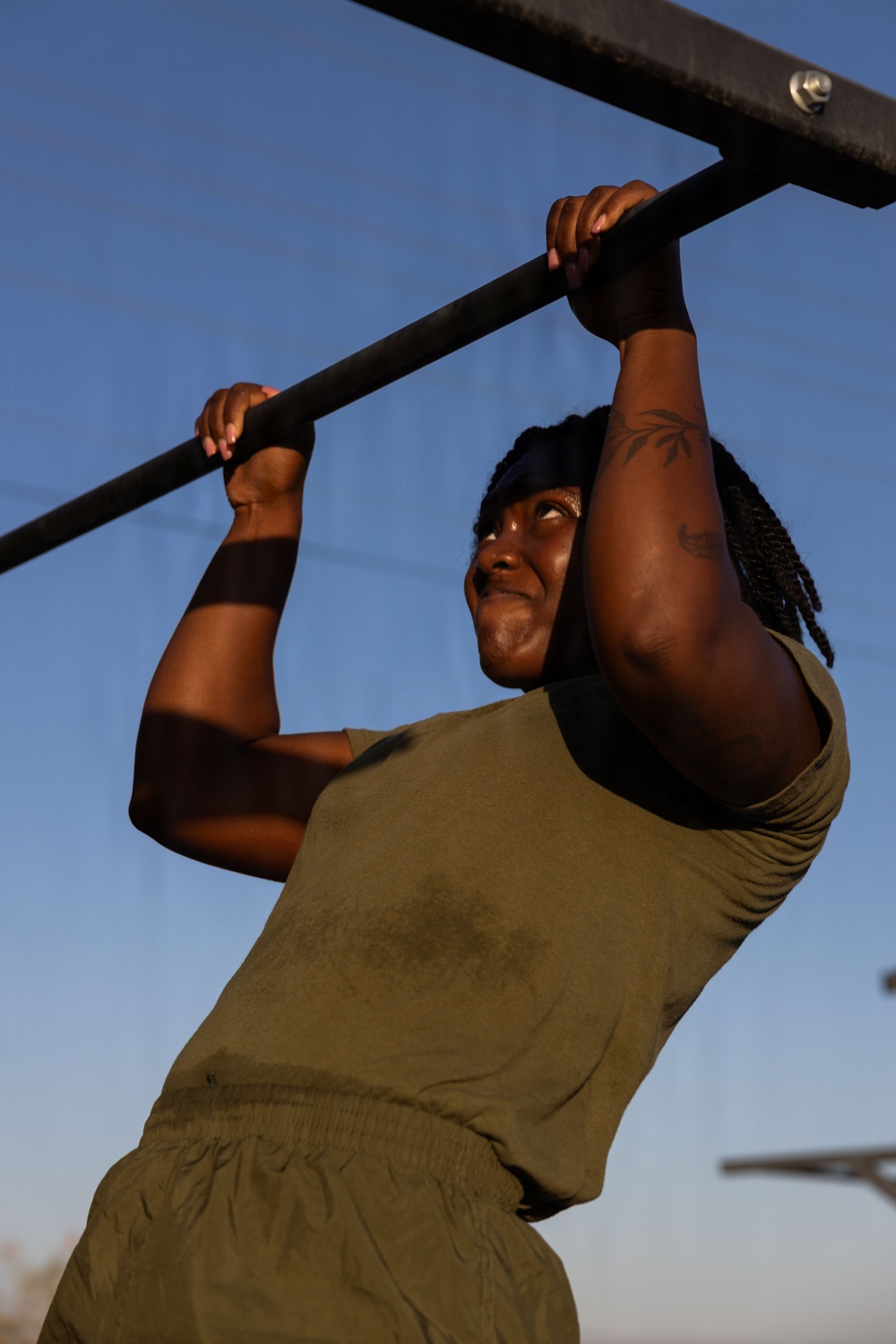 A person is performing pull-ups on an outdoor metal bar, photographed from a low angle against a clear blue sky. They are wearing olive/army green athletic clothing and a dark headband, with visible tattoos on their right arm. The image is taken during golden hour lighting, casting warm shadows across the subject.