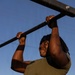 A person is performing pull-ups on an outdoor metal bar, photographed from a low angle against a clear blue sky. They are wearing olive/army green athletic clothing and a dark headband, with visible tattoos on their right arm. The image is taken during golden hour lighting, casting warm shadows across the subject.