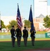 A military color guard of four soldiers in dress uniforms presents the American flag and what appears to be the Indiana state flag on a baseball field during a pre-game ceremony. The ceremony is taking place at a minor league baseball stadium with visible advertising signage including AAA Insurance and NUTRL. Spectators can be seen in the background along the outfield area.
