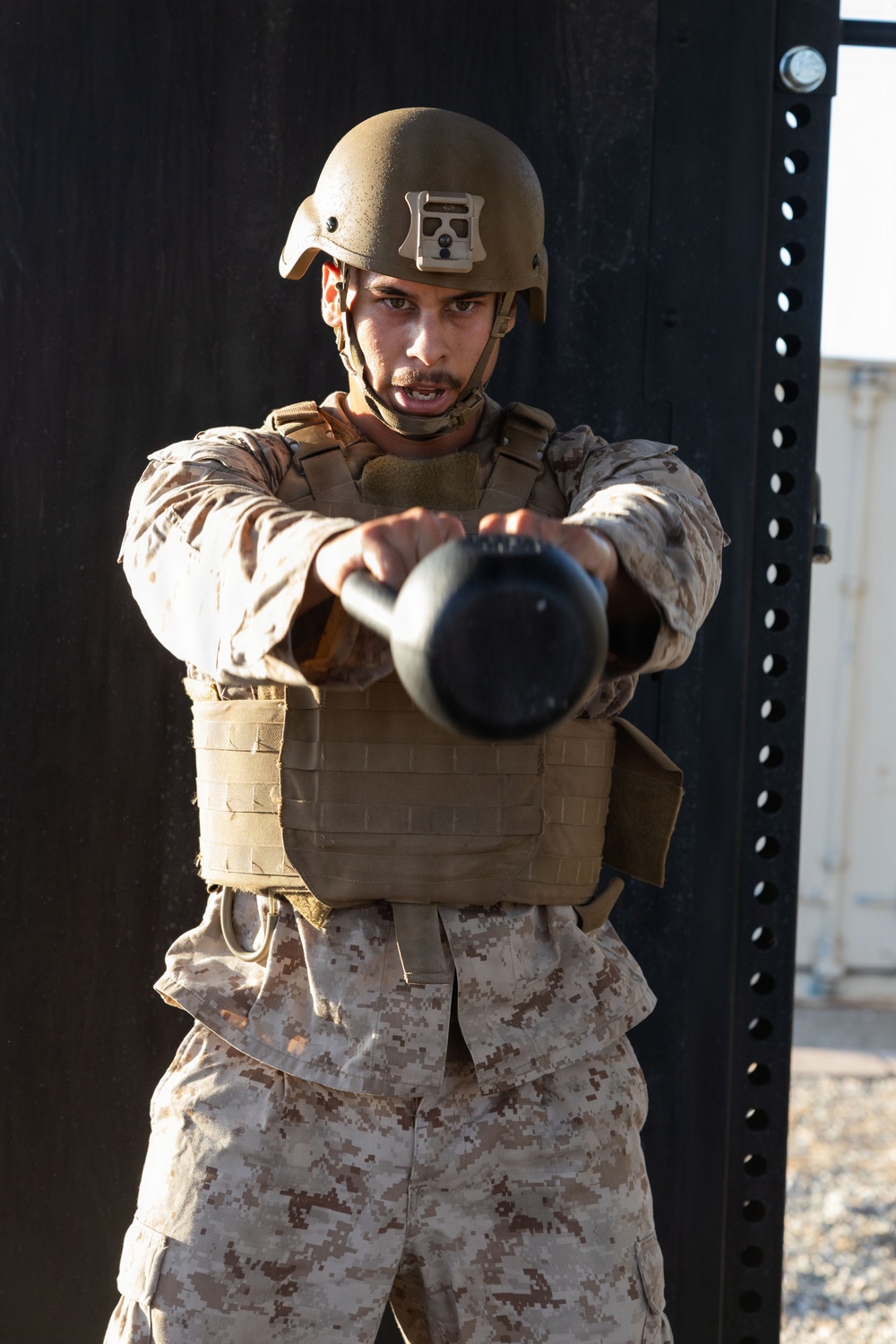 A male service member in full combat gear performs a kettlebell exercise outdoors, extending the weight forward with both arms. He is wearing MARPAT desert camouflage uniform, a tan tactical plate carrier vest, and a ballistic helmet with a mounted device. The image is taken in what appears to be an outdoor fitness or training area.