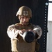 A male service member in full combat gear performs a kettlebell exercise outdoors, extending the weight forward with both arms. He is wearing MARPAT desert camouflage uniform, a tan tactical plate carrier vest, and a ballistic helmet with a mounted device. The image is taken in what appears to be an outdoor fitness or training area.