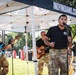 A military band performs outdoors at what appears to be a public event under a canopy tent branded with 'INDYINDIANS.COM'. Three visible performers wear military camouflage (OCP/MultiCam) uniforms, with a lead vocalist at a microphone in the foreground and two guitarists behind him. The event appears to take place in a park or outdoor venue during daytime.