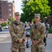 Two U.S. Army soldiers in OCP (Operational Camouflage Pattern) uniforms are standing together on a sidewalk in what appears to be an urban setting. The soldier on the left is identified by name tape as 'ZOLLER' with Staff Sergeant rank insignia and a '1st Infantry Division' patch, while the soldier on the right is identified as 'MCLIN' with Sergeant rank and an 'MP' (Military Police) brassard. Both appear to be on duty at a public event.