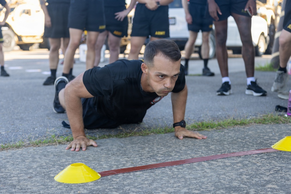 A male soldier in black Army PT (Physical Training) uniform is performing push-ups on a paved surface, observed by a group of fellow soldiers standing in the background. Yellow marker cones and a red/brown strap are visible on the ground, suggesting a structured fitness test or training exercise. The setting appears to be an outdoor military training area with vehicles visible in the background.