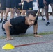 A male soldier in black Army PT (Physical Training) uniform is performing push-ups on a paved surface, observed by a group of fellow soldiers standing in the background. Yellow marker cones and a red/brown strap are visible on the ground, suggesting a structured fitness test or training exercise. The setting appears to be an outdoor military training area with vehicles visible in the background.