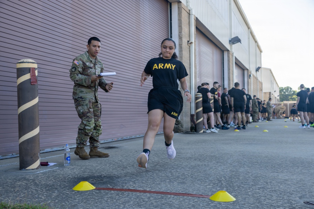 A U.S. Army soldier in PT (Physical Training) uniform is performing a sprint or agility drill on a paved surface near a building with roll-up garage doors. A uniformed soldier in OCP (Operational Camouflage Pattern) is observing and recording results with a clipboard. A group of additional soldiers in PT gear are lined up in the background awaiting their turn.