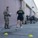 A U.S. Army soldier in PT (Physical Training) uniform is performing a sprint or agility drill on a paved surface near a building with roll-up garage doors. A uniformed soldier in OCP (Operational Camouflage Pattern) is observing and recording results with a clipboard. A group of additional soldiers in PT gear are lined up in the background awaiting their turn.