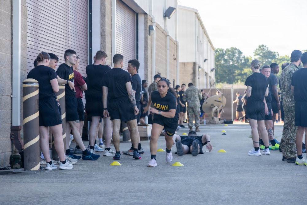 A group of U.S. Army soldiers in black PT (physical training) uniforms are participating in an outdoor fitness exercise or assessment near a military building. One female soldier is prominently running or performing an agility drill through yellow cone markers on the pavement. Other soldiers are lined up along the building wall, waiting their turn or observing.