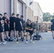 A group of U.S. Army soldiers in black PT (physical training) uniforms are participating in an outdoor fitness exercise or assessment near a military building. One female soldier is prominently running or performing an agility drill through yellow cone markers on the pavement. Other soldiers are lined up along the building wall, waiting their turn or observing.
