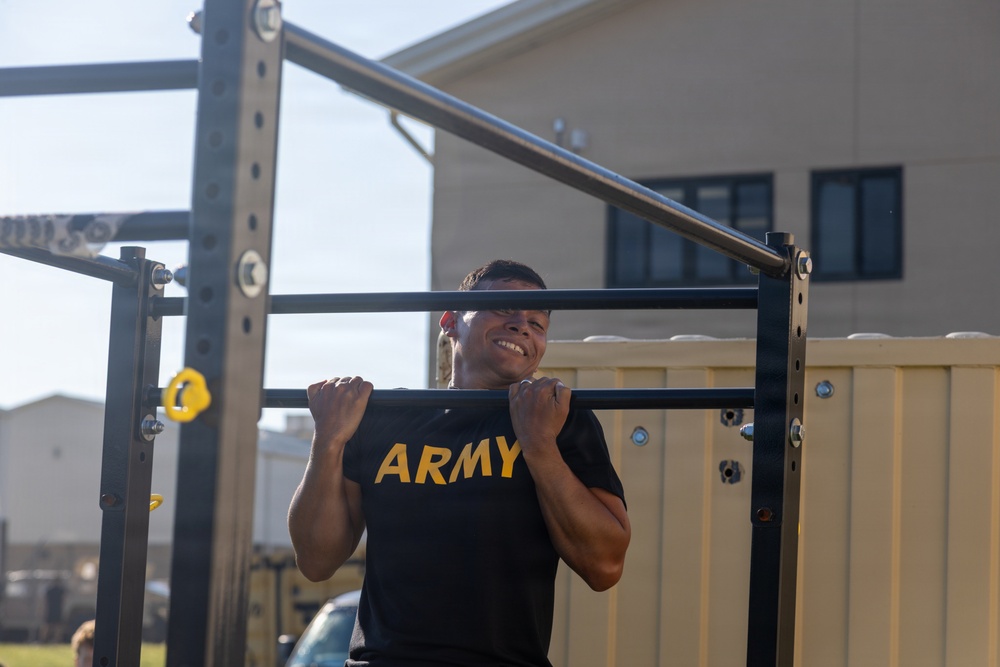 A soldier wearing a black U.S. Army physical training shirt is performing pull-ups on an outdoor fitness rack. The individual is smiling while executing the exercise, with their chin at or above the bar level. The setting appears to be a military installation with institutional buildings visible in the background.