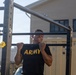 A soldier wearing a black U.S. Army physical training shirt is performing pull-ups on an outdoor fitness rack. The individual is smiling while executing the exercise, with their chin at or above the bar level. The setting appears to be a military installation with institutional buildings visible in the background.